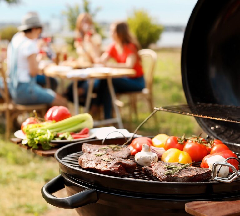 Bunte Gemüse und Steaks werden auf einem Grill zubereitet, während eine Gruppe von Freunden im Hintergrund lacht und isst. | © Adobe Stock/New Africa