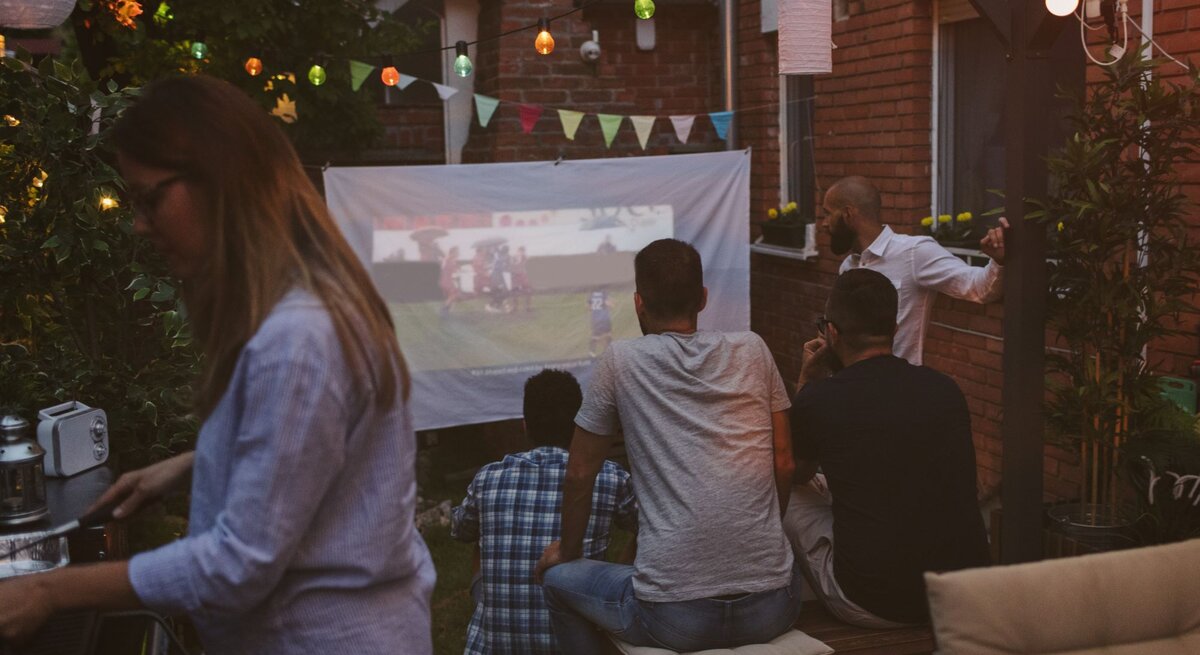 Eine Gruppe von Männern schaut am Abend im Garten ein Fußballspiel auf einer Leinwand, während eine Frau am Grill steht.