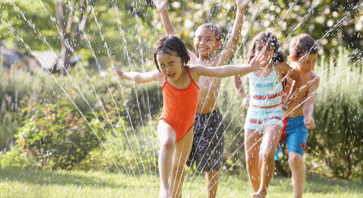 Vier Kinder in bunter Badebekleidung rennen lachend durch Wasserstrahlen eines Rasensprengers auf einer grünen Wiese an einem sonnigen Tag.