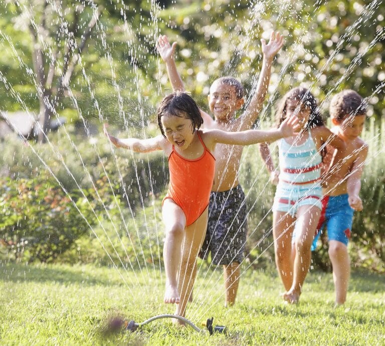 Vier Kinder in bunter Badebekleidung rennen lachend durch Wasserstrahlen eines Rasensprengers auf einer grünen Wiese an einem sonnigen Tag. | © Getty Images/Ariel Skelley