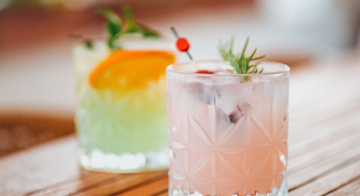 Zwei erfrischende Cocktails in Kristallgläsern auf einem Holztisch, der vordere rosa mit Rosmarin und Beeren, der hintere grün mit Orangenscheibe und Minze.