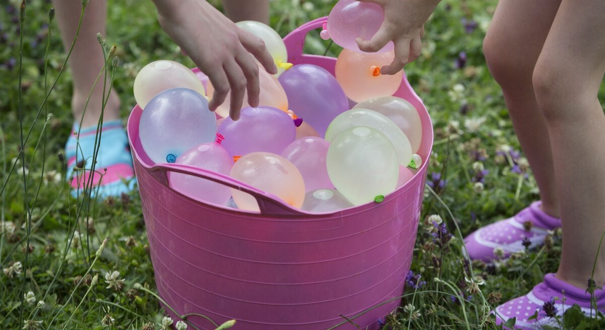 Zwei Kinder beugen sich über einen rosa Eimer voller bunter Wasserbomben auf einer grünen Wiese.