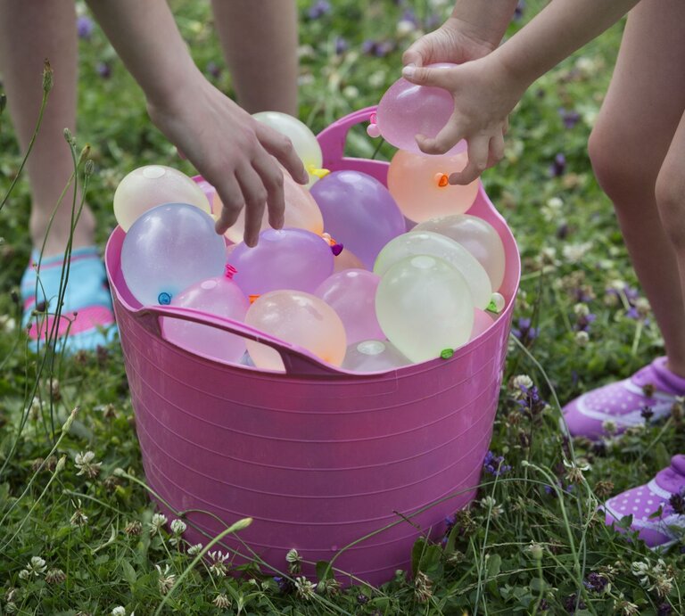 Zwei Kinder beugen sich über einen rosa Eimer voller bunter Wasserbomben auf einer grünen Wiese. | © Getty Images/Elva Etienne