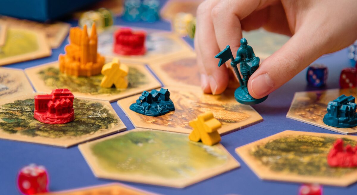 Eine Hand platziert eine blaue Spielfigur mit Fahne auf ein sechseckiges Spielfeld mit bunten Gebäuden und Würfeln.