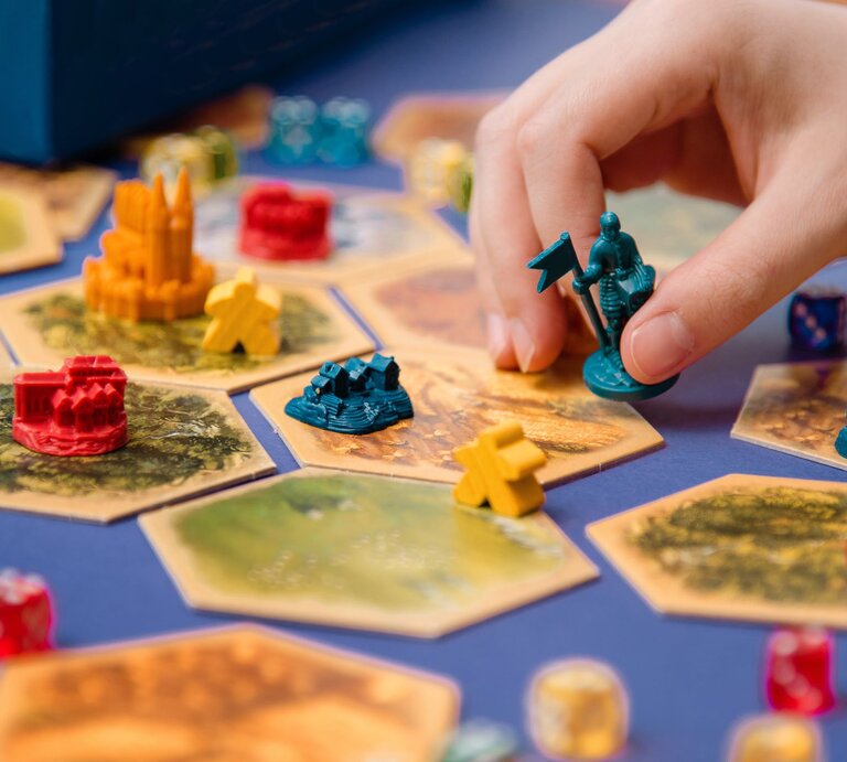 Eine Hand platziert eine blaue Spielfigur mit Fahne auf ein sechseckiges Spielfeld mit bunten Gebäuden und Würfeln. | © Adobe Stock/Alena Gerasimova