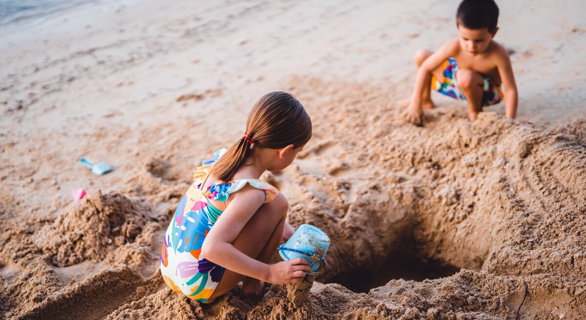 Zwei junge Kinder in farbenfroher Strandkleidung buddeln im Sand an einem leeren Strand ein Loch.
