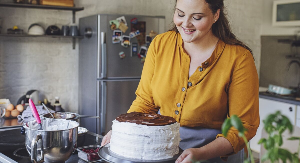 Junge Frau mit braunen Haaren und gelber Bluse dekoriert in einer modernen Küche lächelnd eine selbst gemachte Sahnetorte.