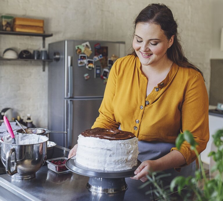 Junge Frau mit braunen Haaren und gelber Bluse dekoriert in einer modernen Küche lächelnd eine selbst gemachte Sahnetorte. | © Getty Images/miniseries