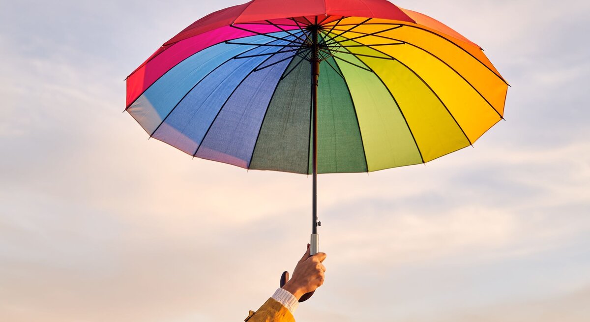 Hand in gelber Jacke hält einen bunten Regenschirm vor einem pastellfarbenen Himmel am Abend.