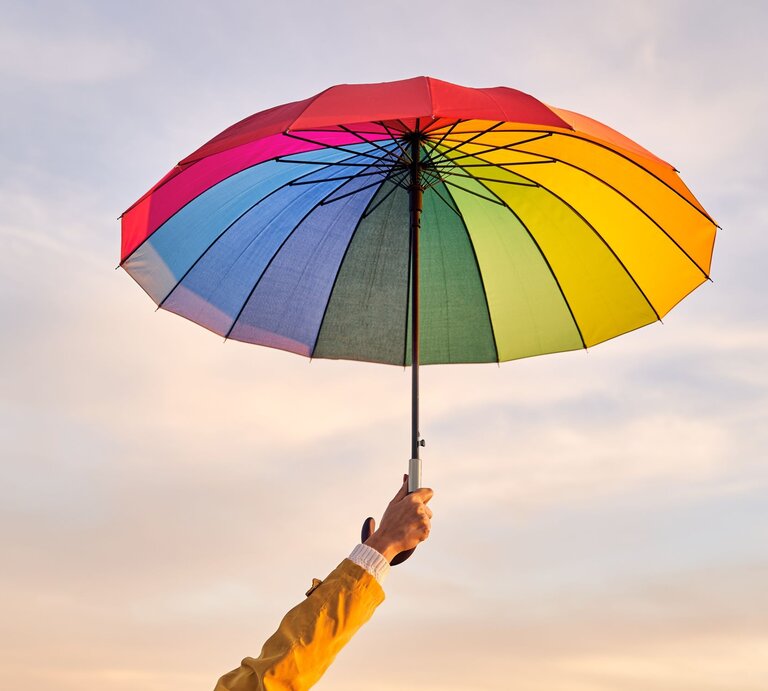 Hand in gelber Jacke hält einen bunten Regenschirm vor einem pastellfarbenen Himmel am Abend. | © Getty Images/Alvaro Medina Jurado