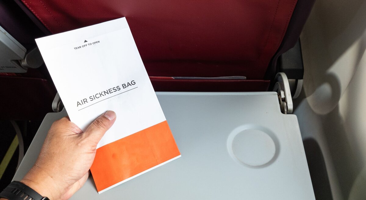 Eine Hand hält einen Spuckbeutel über einem heruntergeklappten Tablett in einem Flugzeug mit dunkelrotem Rücksitz.
