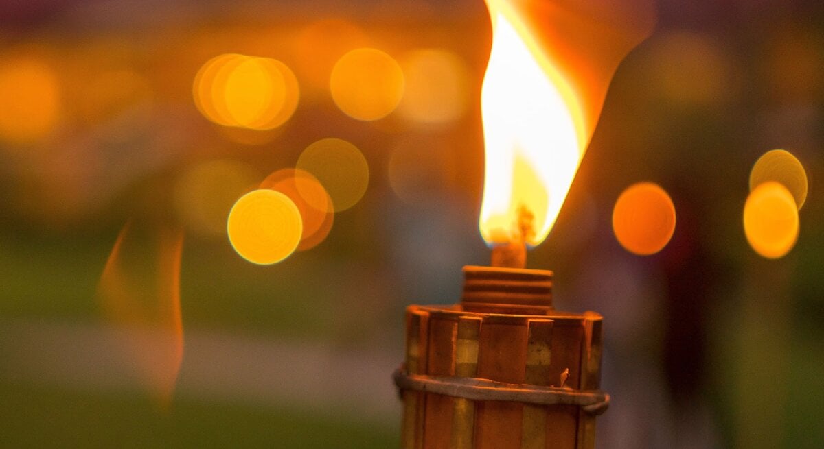 Eine brennende Fackel im Vordergrund mit unscharfen, warmen Lichtern im Hintergrund in der Abenddämmerung.
