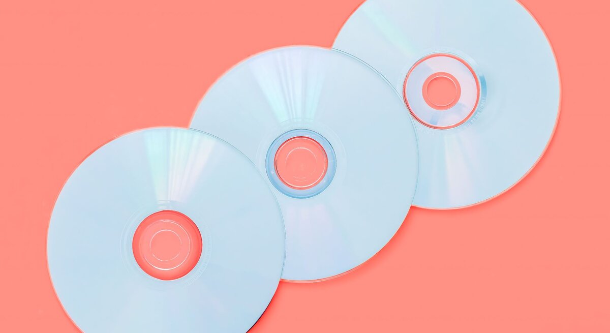 Drei silberne CDs liegen auf einem korallenroten Hintergrund, mit leichtem Schimmer von Lichtreflexen.