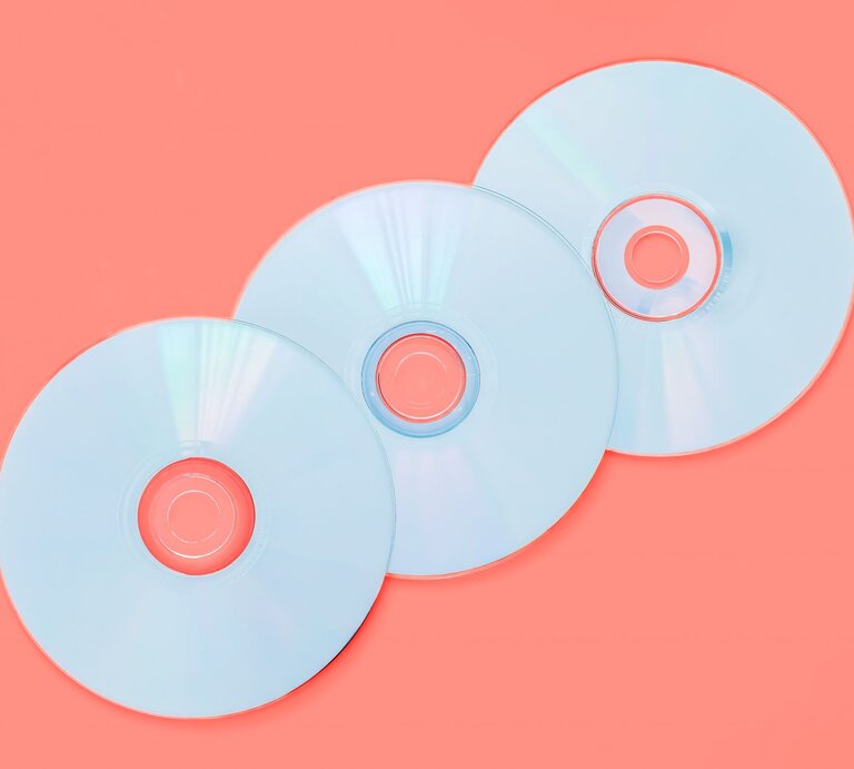 Drei silberne CDs liegen auf einem korallenroten Hintergrund, mit leichtem Schimmer von Lichtreflexen. | © GettyImages/Grace Cary