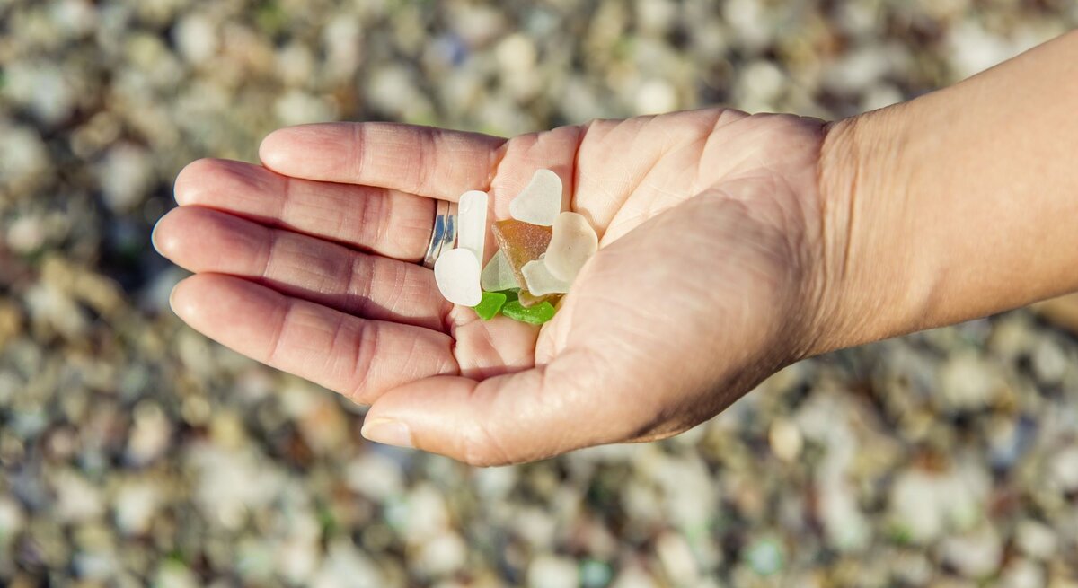 Eine Hand mit silbernem Ring hält bunte Glasstücke auf einem kieseligen Strand.
