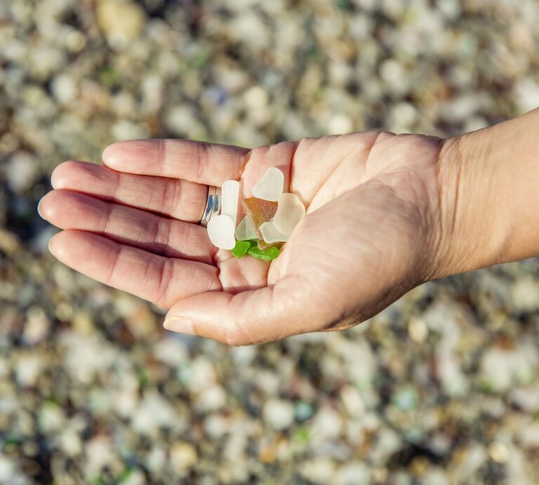 Eine Hand mit silbernem Ring hält bunte Glasstücke auf einem kieseligen Strand. | © GettyImages/JasonDoiy
