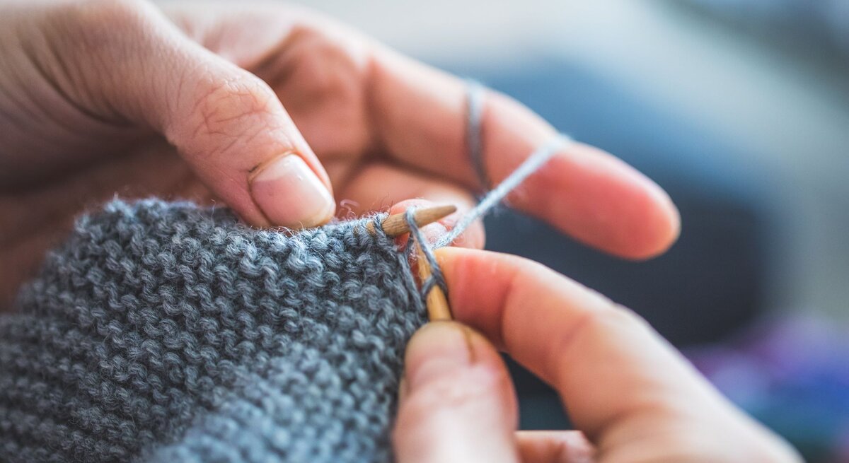 Detailaufnahme von Händen, die geschickt graue Wolle mit einer Stricknadel zu einem Schal verarbeiten.
