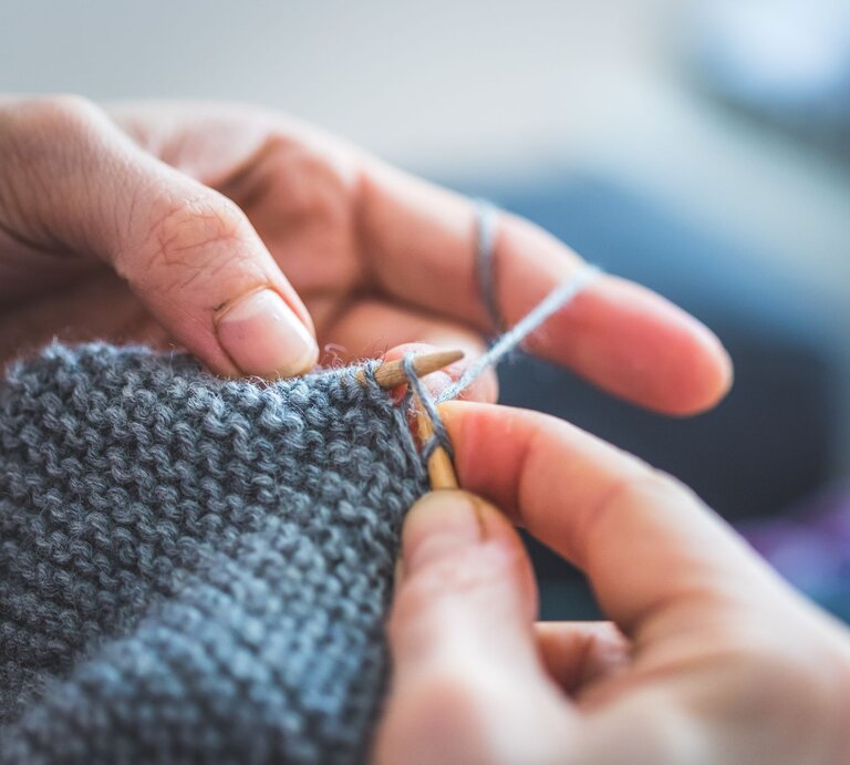 Detailaufnahme von Händen, die geschickt graue Wolle mit einer Stricknadel zu einem Schal verarbeiten. | © Adobe Stock/ Patrick Daxenbichler