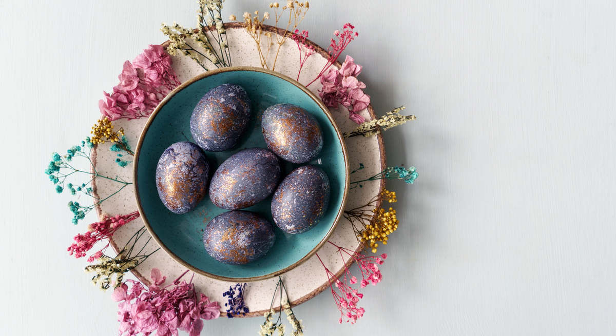 Das Bild zeigt einen liebevoll arrangierten Teller mit marmorierten Ostereiern, umgeben von getrockneten Blumen verschiedener Farben. Die Eier präsentieren sich in einer tiefblauen Farbe mit metallisch wirkenden, kupferfarbenen Sprenkeln, die eine kunstvolle und marmorierte Oberfläche erzeugen. Der Teller steht auf einem größeren, zweifarbigen Teller, der als Unterlage dient und das Arrangement abrundet. Der Hintergrund ist hell und neutral, was die Farbvielfalt der Eier und Blumen hervorhebt und eine ruhige, ästhetische Atmosphäre schafft.