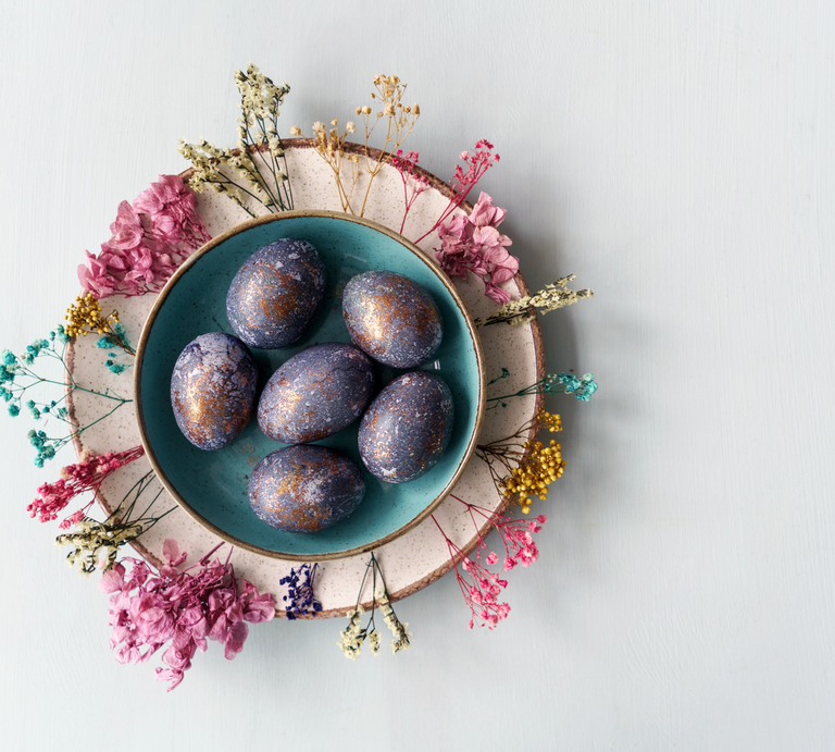 Das Bild zeigt einen liebevoll arrangierten Teller mit marmorierten Ostereiern, umgeben von getrockneten Blumen verschiedener Farben. Die Eier präsentieren sich in einer tiefblauen Farbe mit metallisch wirkenden, kupferfarbenen Sprenkeln, die eine kunstvolle und marmorierte Oberfläche erzeugen. Der Teller steht auf einem größeren, zweifarbigen Teller, der als Unterlage dient und das Arrangement abrundet. Der Hintergrund ist hell und neutral, was die Farbvielfalt der Eier und Blumen hervorhebt und eine ruhige, ästhetische Atmosphäre schafft. | © GettyImages/Tatyana Maximova