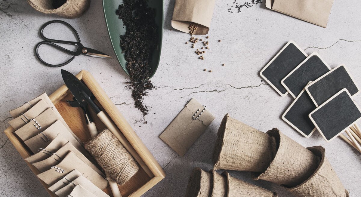  Dieses Bild zeigt Gartenbedarf und Utensilien, die wahrscheinlich für die Aussaat und Anzucht von Pflanzen verwendet werden. Zu sehen sind biologisch abbaubare Anzuchttöpfe aus Papier, die zur Keimung von Samen bevor sie ins Freiland gepflanzt werden, benutzt werden können. Außerdem gibt es eine Schale mit Erde, kleine Papiertüten, die vermutlich Samen enthalten, sowie eine Bambusbox mit Gartengeräten wie einer kleinen Schaufel und einer Harke. Ebenfalls abgebildet sind Gartenetiketten aus Schiefer, die wahrscheinlich zur Beschriftung der Pflanzen dienen, sowie eine Garnrolle, die zum Binden der Pflanzen oder Markieren von Reihen verwendet werden kann. Das Arrangement auf einer strukturierten, hellen Oberfläche vermittelt eine organisierte und naturnahe Gartenarbeitsszene.