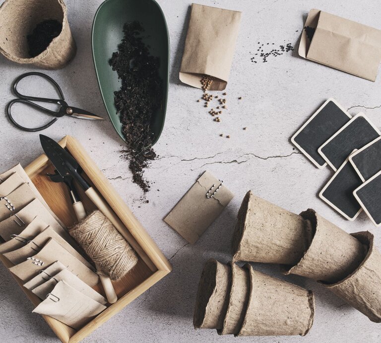  Dieses Bild zeigt Gartenbedarf und Utensilien, die wahrscheinlich für die Aussaat und Anzucht von Pflanzen verwendet werden. Zu sehen sind biologisch abbaubare Anzuchttöpfe aus Papier, die zur Keimung von Samen bevor sie ins Freiland gepflanzt werden, benutzt werden können. Außerdem gibt es eine Schale mit Erde, kleine Papiertüten, die vermutlich Samen enthalten, sowie eine Bambusbox mit Gartengeräten wie einer kleinen Schaufel und einer Harke. Ebenfalls abgebildet sind Gartenetiketten aus Schiefer, die wahrscheinlich zur Beschriftung der Pflanzen dienen, sowie eine Garnrolle, die zum Binden der Pflanzen oder Markieren von Reihen verwendet werden kann. Das Arrangement auf einer strukturierten, hellen Oberfläche vermittelt eine organisierte und naturnahe Gartenarbeitsszene. | © Gettyimages/Mkovalevskaya