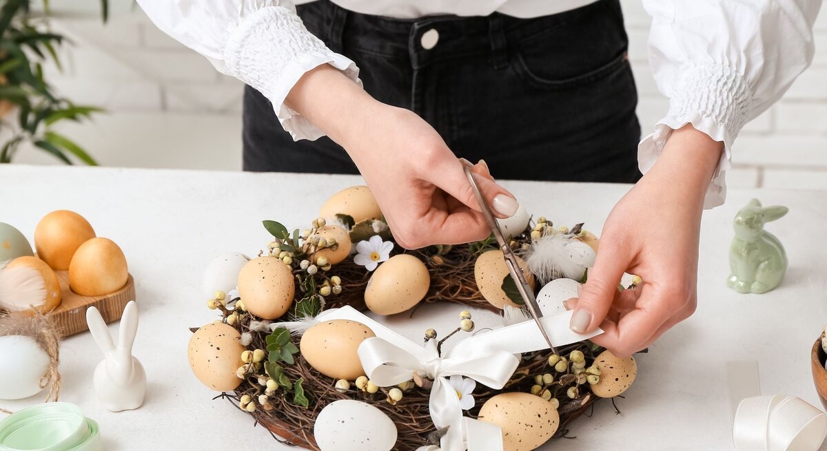 Junge Frau mit weißer Bluse bindet ein weißes Band an einen Osterkranz aus Zweigen, Eiern, Blüten und Beeren auf weißem Tisch.