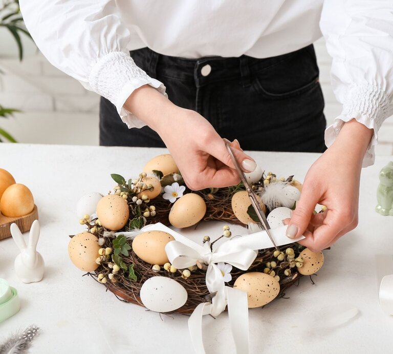 Junge Frau mit weißer Bluse bindet ein weißes Band an einen Osterkranz aus Zweigen, Eiern, Blüten und Beeren auf weißem Tisch. | © Shutterstock/ Pixel-Shot