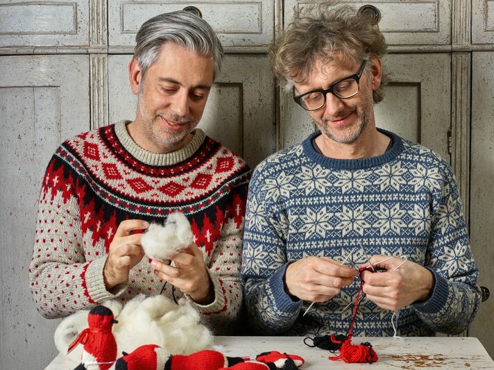 Zwei Männer, die gemeinsam Handarbeiten ausführen. Der Mann links im Bild trägt einen rot-schwarz-weißen Weihnachtspullover, der Mann rechts einen blau-weißen Pullover im skandinavischen Stil. Beide Männer haben graue Haare und scheinen in ihrer Handarbeit vertieft zu sein. Auf dem Tisch vor ihnen liegen fertige gestrickte Objekte. Sie sitzen vor einem Schrank mit antikem Aussehen | © Topp Verlag
