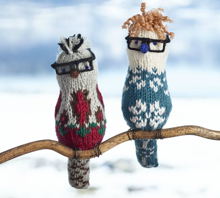 Zwei gestrickte Vogelfiguren auf einem Ast: Die linke hat ein rotes Gestrick mit grünen und weißen Mustern, trägt eine schwarze Brille und hat eine kleine gestrickte Nase. Die rechte hat ein weiß-blau gestricktes Muster, trägt ebenfalls eine schwarze Brille und hat lockiges, braunes Haar oben auf dem Kopf. Im Hintergrund ist verschwommen eine schneebedeckte Landschaft zu erkennen. | © Topp Verlag