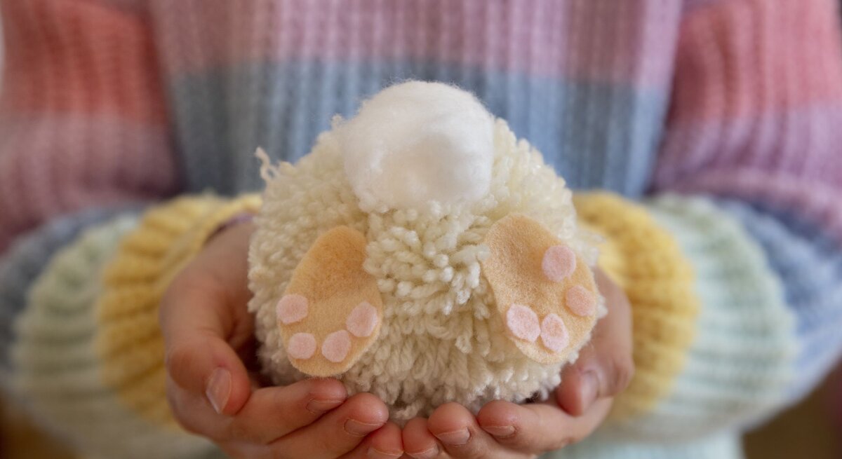  Das Bild zeigt ein handgefertigtes Osterdekorationsobjekt aus Wolle, das von zwei Händen gehalten wird. Die Hände sind um ein wolliges, rundes Objekt geschlossen, das scheinbar einen kleinen Osterhasen darstellt, komplett mit weißen und rosa farbenen Woll-Details, die die Pfoten und die Fußballen abbilden. Oben auf dem Objekt befindet sich eine kleinere weiße Wollkugel, die vermutlich den Schwanz des Hasen repräsentiert. Die Person trägt einen gestreiften Pullover in Pastelltönen, was eine weiche und warme Frühlingsstimmung vermittelt. Der Fokus auf das wollige Osterhasenobjekt in den sorgsam haltenden Händen unterstreicht die liebevolle und sorgfältige Herstellung von saisonaler Handwerkskunst.