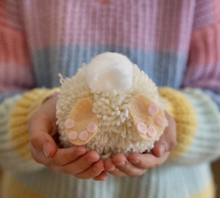  Das Bild zeigt ein handgefertigtes Osterdekorationsobjekt aus Wolle, das von zwei Händen gehalten wird. Die Hände sind um ein wolliges, rundes Objekt geschlossen, das scheinbar einen kleinen Osterhasen darstellt, komplett mit weißen und rosa farbenen Woll-Details, die die Pfoten und die Fußballen abbilden. Oben auf dem Objekt befindet sich eine kleinere weiße Wollkugel, die vermutlich den Schwanz des Hasen repräsentiert. Die Person trägt einen gestreiften Pullover in Pastelltönen, was eine weiche und warme Frühlingsstimmung vermittelt. Der Fokus auf das wollige Osterhasenobjekt in den sorgsam haltenden Händen unterstreicht die liebevolle und sorgfältige Herstellung von saisonaler Handwerkskunst. | © gettyimages/	Elva Etienne