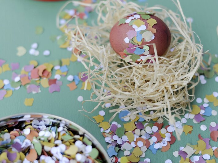  Das Bild zeigt ein sorgfältig dekoriertes Osterei, das in einem Nest aus natürlich aussehendem Stroh platziert ist. Das Ei ist mit bunten Konfettistücken verziert, die auf seiner Oberfläche haften und ihm ein festliches Aussehen verleihen. | © gettyimages/Westend61