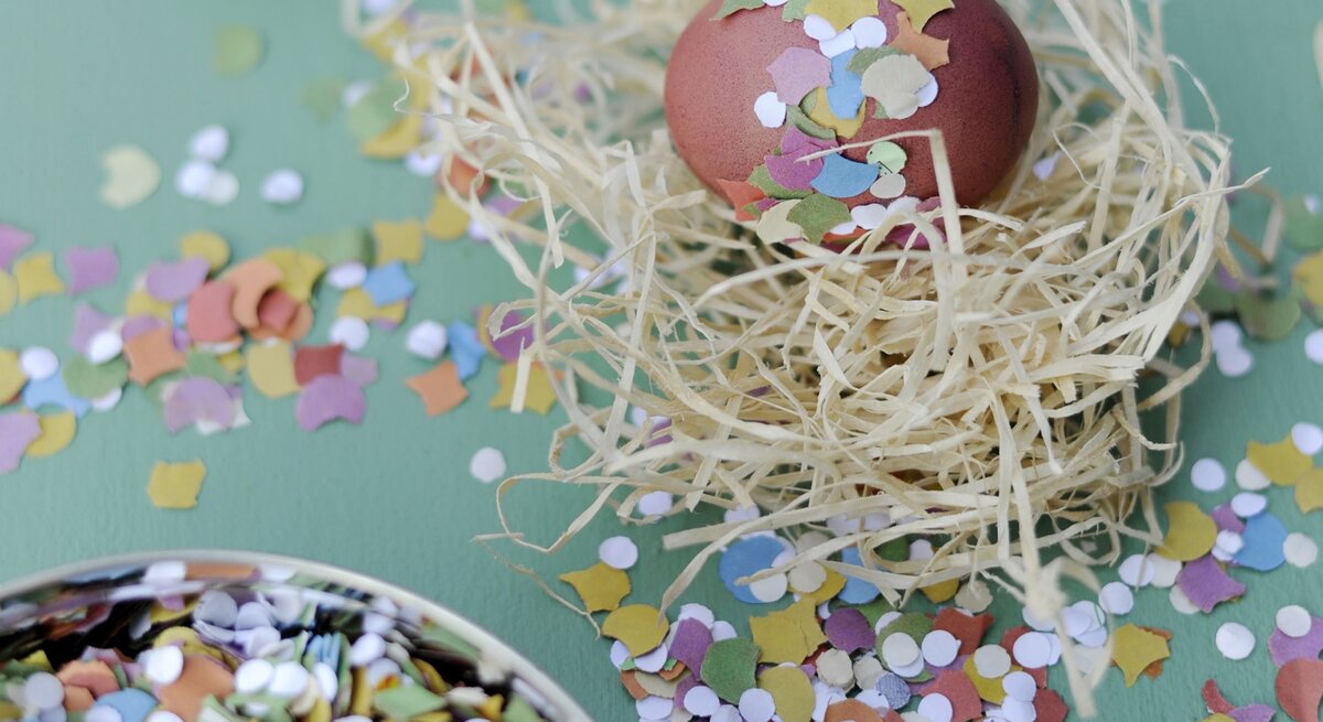  Das Bild zeigt ein sorgfältig dekoriertes Osterei, das in einem Nest aus natürlich aussehendem Stroh platziert ist. Das Ei ist mit bunten Konfettistücken verziert, die auf seiner Oberfläche haften und ihm ein festliches Aussehen verleihen.