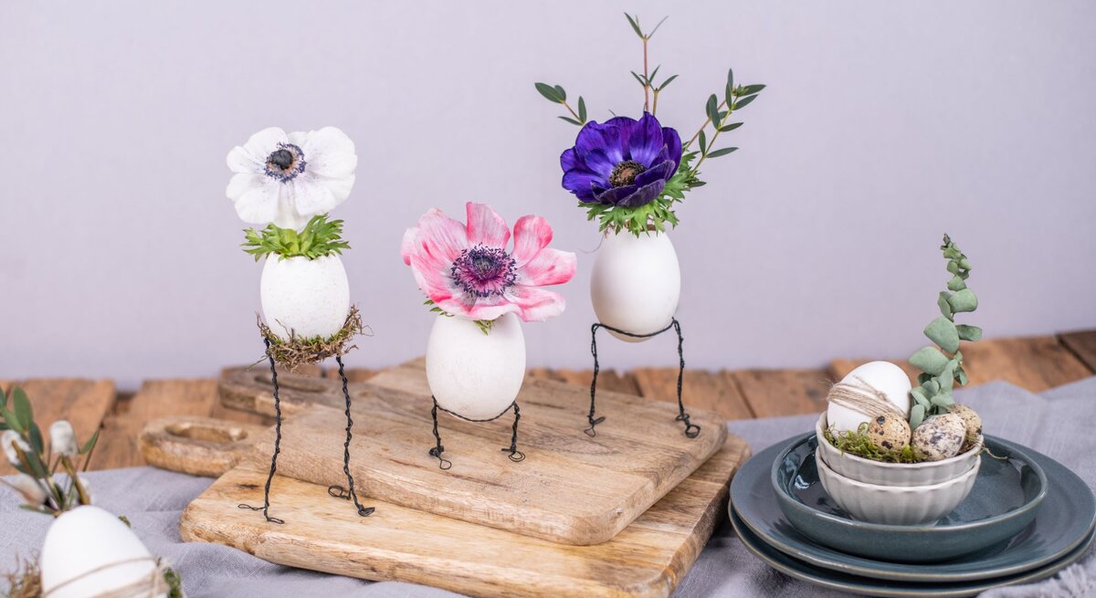  Drei dekorative Eier auf Drahtbeinen mit aufgesetzten Blumenköpfen – ein weißes mit einer Anemone, ein zweites mit einer rosa Mohnblume und ein drittes mit einer dunkelvioletten Anemone – arrangiert auf einem hölzernen Schneidebrett. Auf dem Brett befinden sich auch weitere dekorierte Eier in einer kleinen Schüssel auf einem Stapel von Keramiktellern. Die Szene ist auf einem grauen Tuch mit lila Wand im Hintergrund drapiert, was einen kreativen Basteltrend mit Draht und Naturmaterialien darstellt.