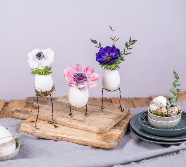  Drei dekorative Eier auf Drahtbeinen mit aufgesetzten Blumenköpfen – ein weißes mit einer Anemone, ein zweites mit einer rosa Mohnblume und ein drittes mit einer dunkelvioletten Anemone – arrangiert auf einem hölzernen Schneidebrett. Auf dem Brett befinden sich auch weitere dekorierte Eier in einer kleinen Schüssel auf einem Stapel von Keramiktellern. Die Szene ist auf einem grauen Tuch mit lila Wand im Hintergrund drapiert, was einen kreativen Basteltrend mit Draht und Naturmaterialien darstellt. | © Adobe Stock/knipseria