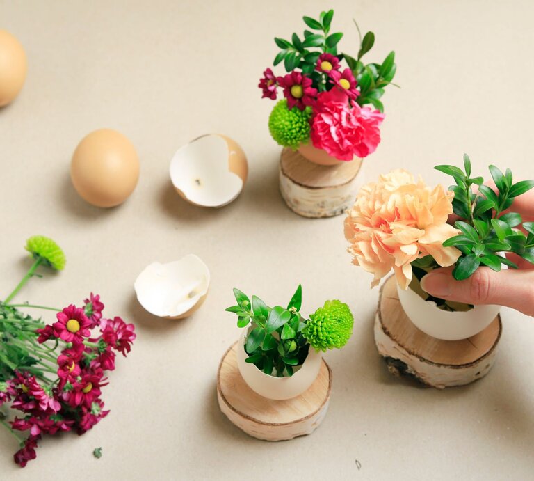 Eine Hand arrangiert eine zarte Blumen-Deko in einer halben Eierschale auf einem Holzsockel, umgeben von Blüten und Eierschalen. | © Shutterstock/ Agnes Kantaruk