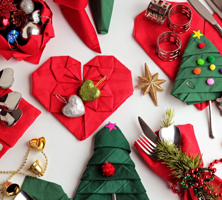 Festliche Tischdekoration mit roten Servietten in Herzform, grünen Servietten als Weihnachtsbäume gefaltet, Kugeln und goldenem Stern. | © GettyImages/Gulsen Ozcan