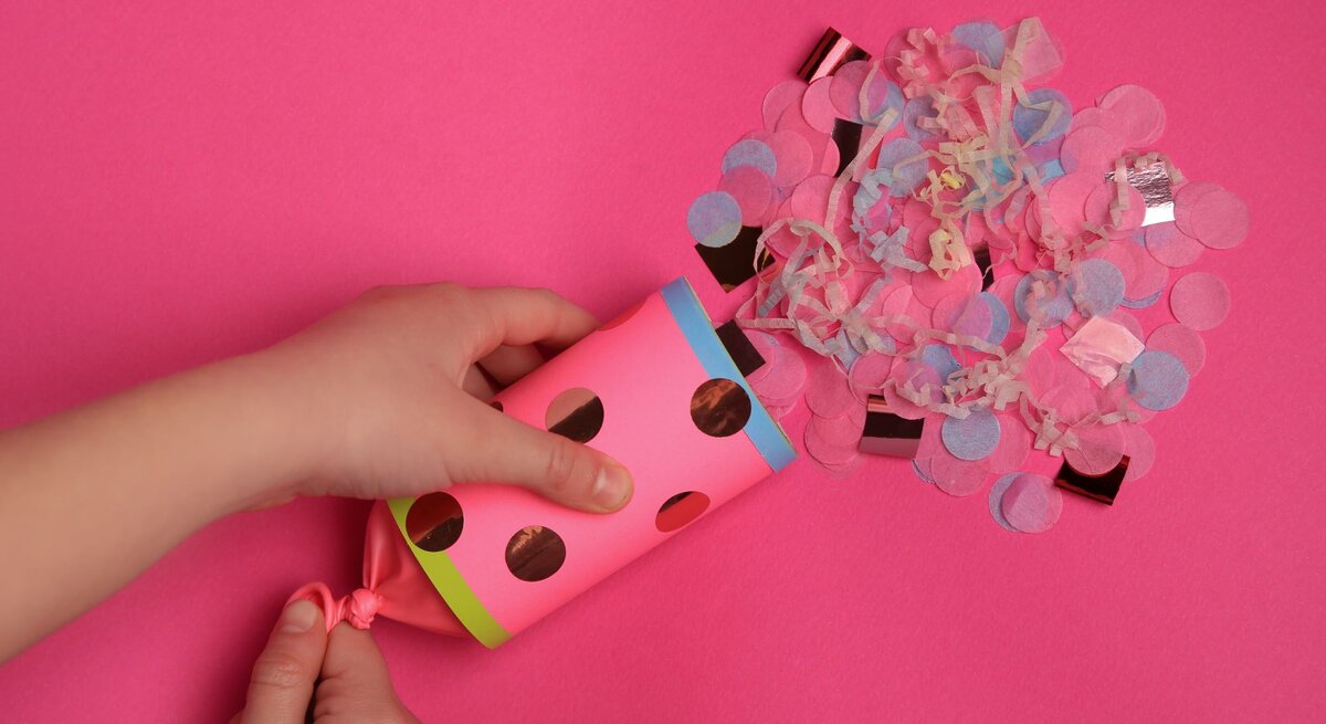 Hände halten eine selbstgebastelte Konfetti-Kanone aus Pappe über einen pinken Hintergrund, aus der gerade buntes Konfetti herausströmt, was eine lebhafte und festliche Atmosphäre erzeugt.