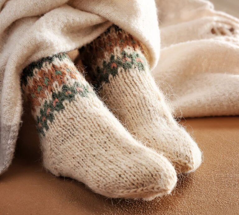 Kuschelige gestrickte Wollsocken mit farbigem Norwegermuster ragen aus beiger Decke, liegend auf einem weichen Sofa in warmem Licht. | © Adobe Stock/ Pixel-Shot
