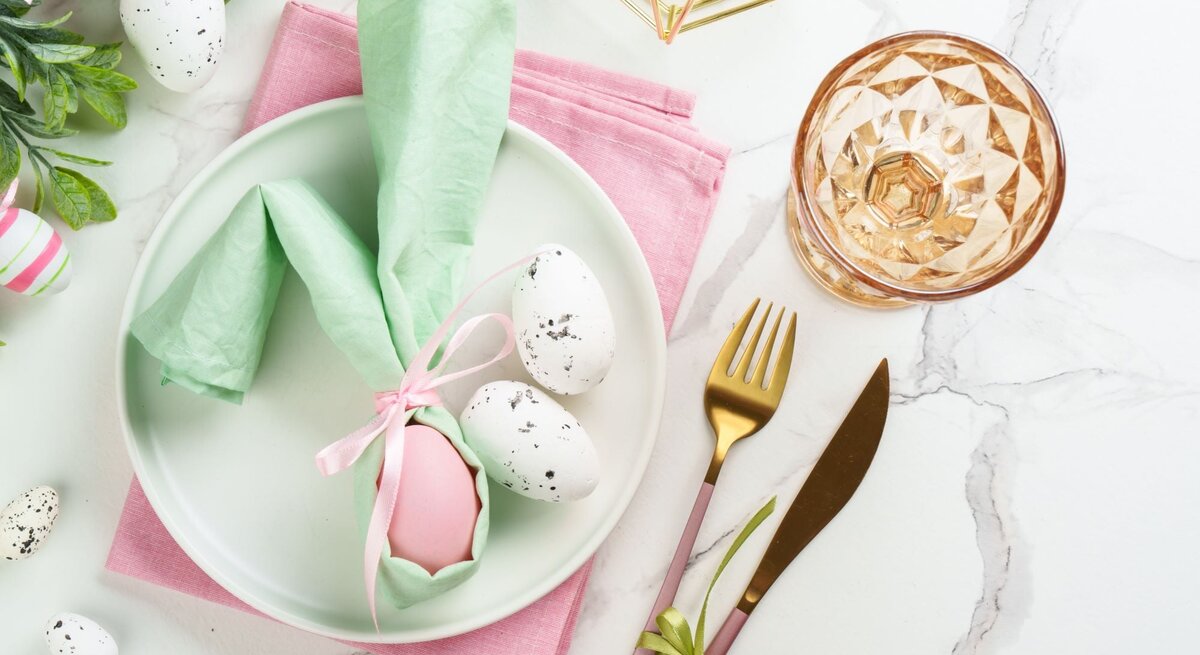 Osterlich gedeckter Tisch mit rosa Serviette, pastellfarbenem Ei im grünen Hasen-Serviettenhalter und goldfarbenem Besteck.