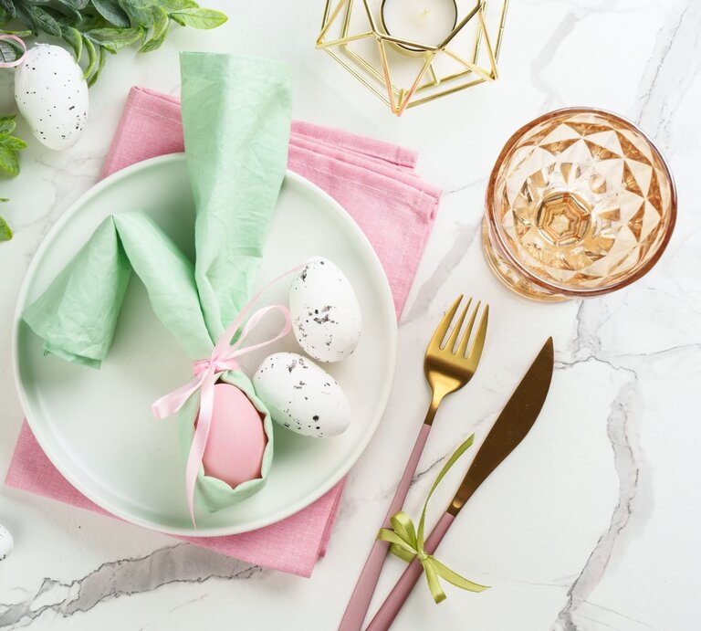 Osterlich gedeckter Tisch mit rosa Serviette, pastellfarbenem Ei im grünen Hasen-Serviettenhalter und goldfarbenem Besteck. | © Shutterstock/ nadianb