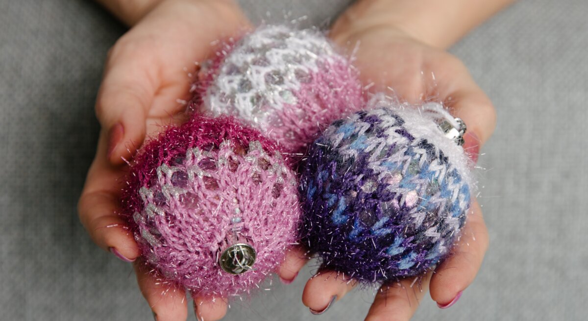 Nahaufnahme von Händen mit rot lackierten Nägeln, die drei gestrickte, glitzernde Weihnachtskugeln in Rosa, Lila und Blau halten.