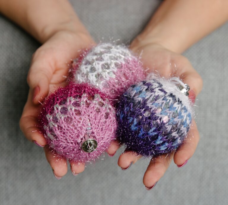 Nahaufnahme von Händen mit rot lackierten Nägeln, die drei gestrickte, glitzernde Weihnachtskugeln in Rosa, Lila und Blau halten. | © Adobe Stock/ AndreyZayats