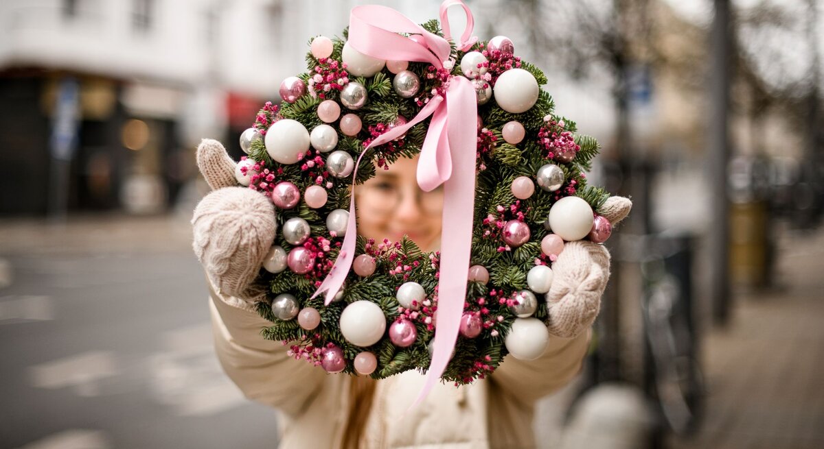 Junge Frau in Winterjacke und Wollhandschuhen hält draußen einen üppigen Adventskranz mit rosa Kugeln und Schleife.