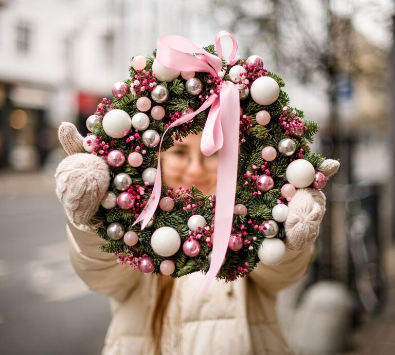Junge Frau in Winterjacke und Wollhandschuhen hält draußen einen üppigen Adventskranz mit rosa Kugeln und Schleife. | © Shutterstock/Maksym Fesenko