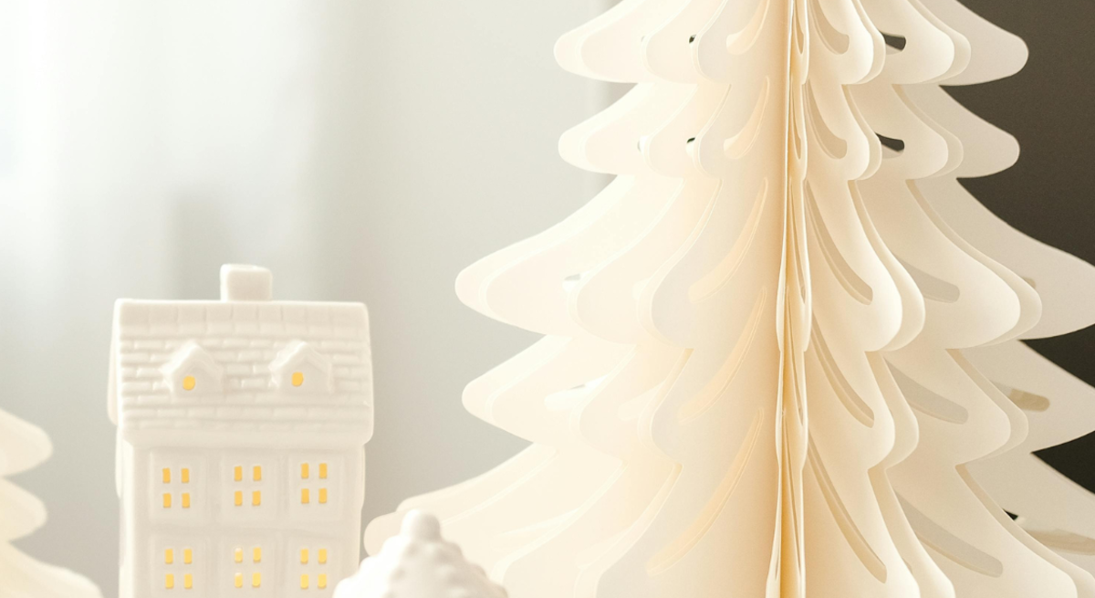 Faltbare Papierbäume in Weiß, Grün und Beige bilden mit leuchtenden Keramikhäuschen ein stimmungsvolles Winterensemble.