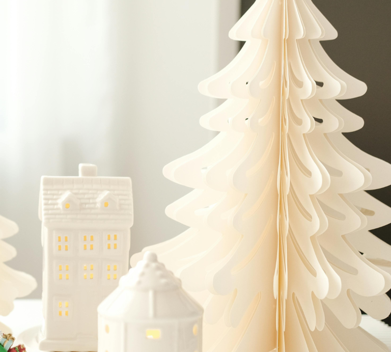 Faltbare Papierbäume in Weiß, Grün und Beige bilden mit leuchtenden Keramikhäuschen ein stimmungsvolles Winterensemble. | © Canva/Collage:bildderfrau.de