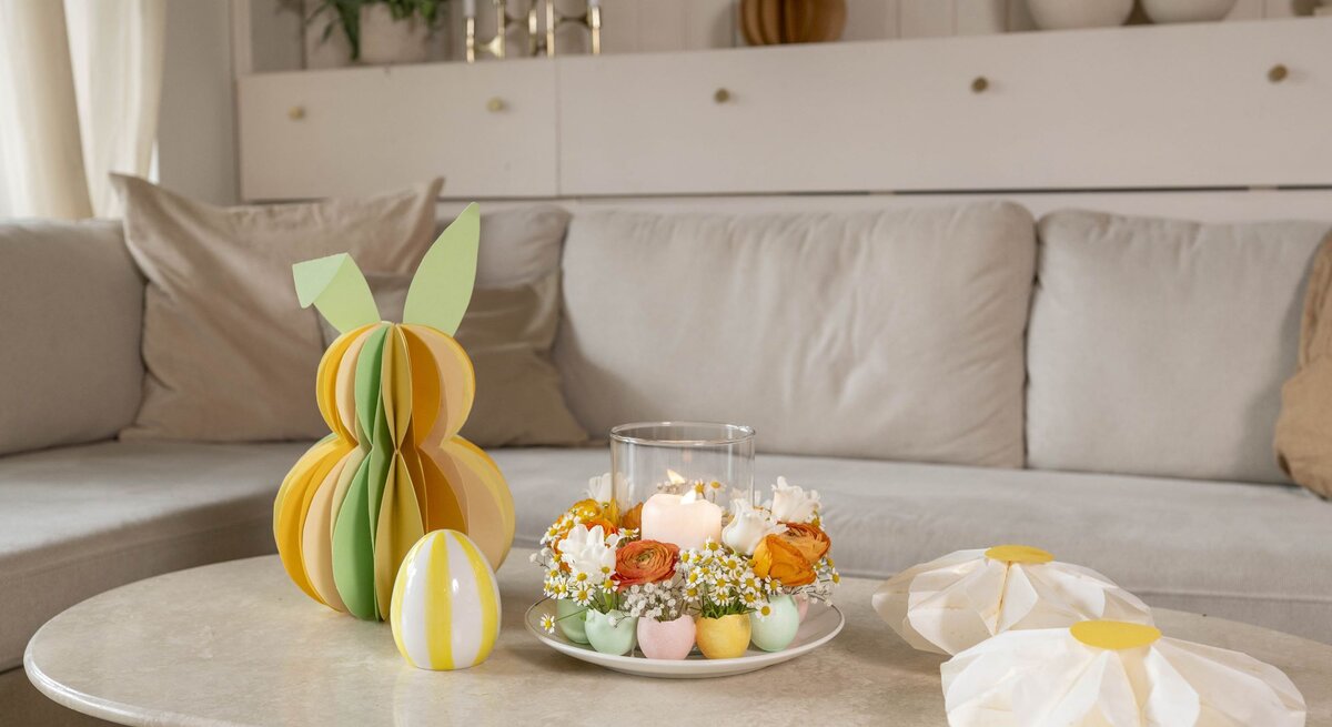 Farbiger Wabenhase aus Papier mit grünen Ohren steht neben Deko-Ei und Osterkranz aus Blumen und Kerzen auf einem Couchtisch.