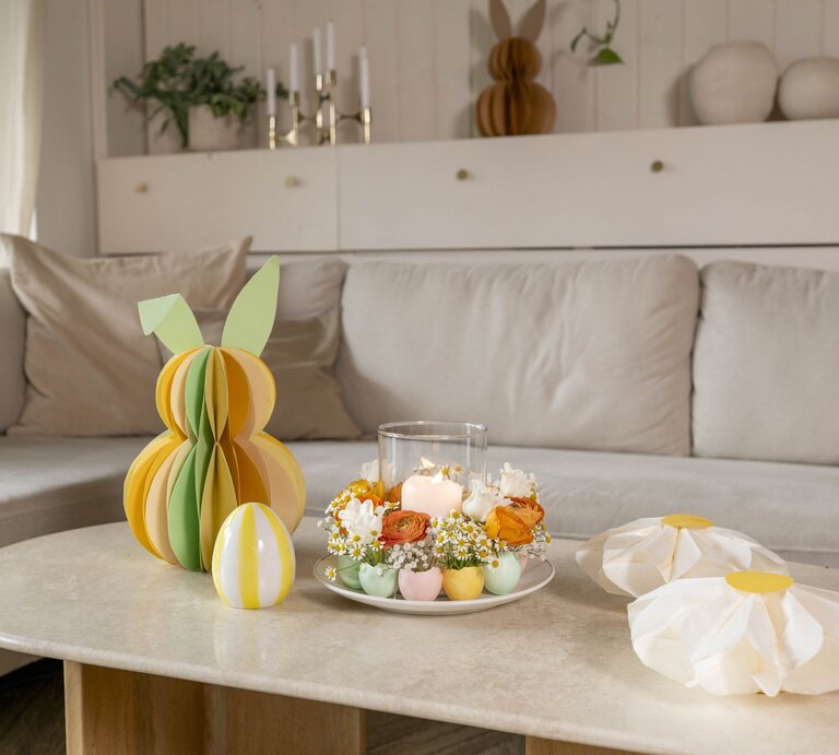 Farbiger Wabenhase aus Papier mit grünen Ohren steht neben Deko-Ei und Osterkranz aus Blumen und Kerzen auf einem Couchtisch. | © BILD der FRAU 